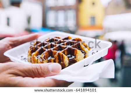 Tourist holds in hand popular street food - Belgium tasty waffle with chocolate sauce on the background of city tourist streets of Bruges, Belgium, Europe. Traditional Belgian dessert, pastry.