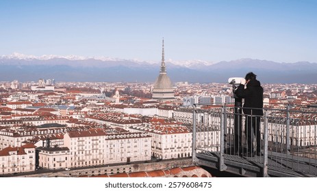 A tourist looks through a sightseeing binoculars on city of Turin (Torino) and Mole Antonelliana. Snowy Alps mountains on background. Piedmont, Italy