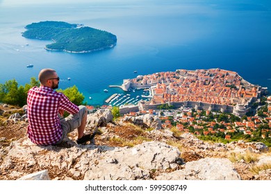 Tourist Man Looking Down To The Old Town Of Dubrovnik, Sitting On The Mountain Above The City, Croatia