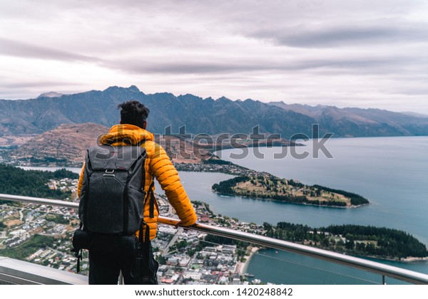 Tourist man looking down at\
Queenstown from Skyline Gondola attraction. View of city from\
viewpoint. Red, yellow. Romantic holiday, travel concepts. New\
Zealand.