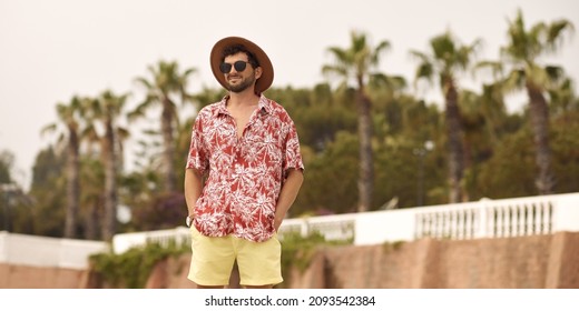 Tourist man on the beach standing outdoors against resort with palm trees in the background during summer holidays. The guy wearing a summer casual shirt hat and sunglasses, standing looking to the - Powered by Shutterstock