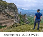 Tourist man standing on cliff edge with view on cliff dwelling house Ypapanti Monastery near Kalambaka seen from opposite hill, Meteora, Thessaly, Greece, Europe. Foggy rainy day mystical atmosphere