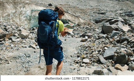 Tourist Man Walks With A Backpack, Studies A Map And Shows A Route In The Mountains Against A Blue Glacier, Slow Motion.