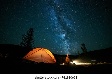 Tourist Near His Camp Tent At Night Under A Sky Full Of Stars. Orange Illuminated Tent.