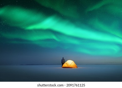 Tourist near yellow tent lighted from the inside against the backdrop of incredible starry sky with Aurora borealis. Amazing night landscape. Northern lights in winter field