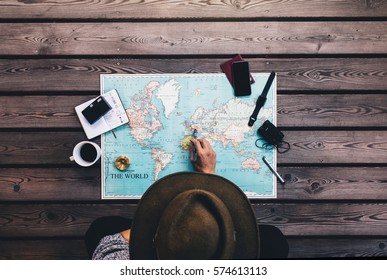 Tourist Pointing At Europe On World Map Surrounded With Binoculars, Compass And Other Travel Accessories. Man Wearing Brown Hat Planning His Tour Looking At The World Map.