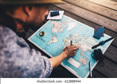 Tourist Pointing At India On World Map Surrounded With Binoculars, Compass And Other Travel Accessories. Man Looking At The World Map Sitting On Wooden Floor.