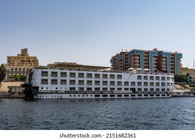 The Tourist Ship Is Moored At The River Bank. Cabin Windows And Railings Are Visible. On The Upper Deck There Is An Awning, Umbrellas. Ripples On The Water. City Houses Against The Blue Sky. Egypt