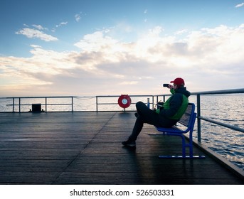 Tourist Sit On Bench On Mole And Take Photos. Man Enjoy Morning At Sea. Sunny Clear Blue Sky, Smooth Water Level