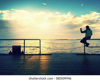 Tourist Sit On Mole Handrail And Takes Pictures. Man Enjoy Morning At Sea. Sunny Clear Blue Sky, Smooth Water Level
