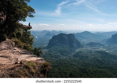 A tourist sits on the cliff edge overhanging the plain on sunny day. Dragon's Crest (Khao Ngon Nak) Viewpoint, Krabi Province, Thailand.