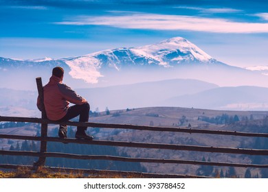 Tourist sitting on a wooden fence and enjoy mountain valley with snow-capped peak on a skynline
