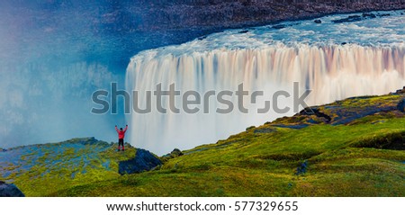 Tourist standing on the shore of falling water of the most powerful waterfall in Europe - Dettifoss with raised hands. Colorful summer sunset in Jokulsargljufur National Park, Iceland. 