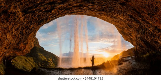 a tourist stands behind the waterfall Seljalandsfoss as the sun sets on a summer evening in southern Iceland - Powered by Shutterstock