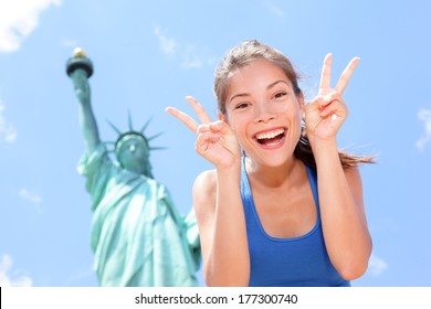 Tourist At Statue Of Liberty, New York, USA Making Funny Face Expression Victory Hand Signs Excited And Happy. Tourism And Travel Concept With Joyful Mixed Race Asian Caucasian Woman.