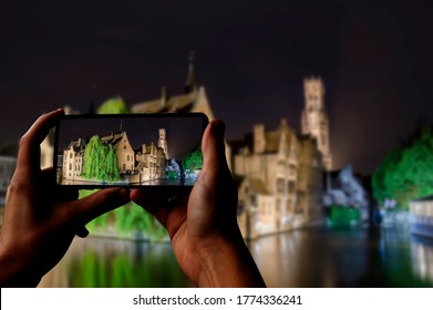 Tourist Taking Photo Of Canal Of Bruges, Tower And Historic Buildings At Night, Bruges, Belgium. Man Holding Phone And Taking Picture.