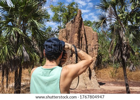 Tourist taking a picture of a big magnetic termite mound. Fit man holding camera,   wearing cap and t-shirt. Strong arms. Litchfield national park, Northern Territory NT, Australia