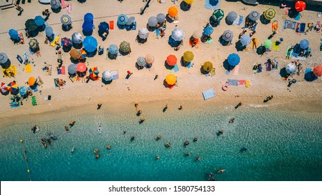 Tourist Under Parasol On Crowded Beach, Beautiful Turquoise And Blue Water, Swimmers, Sunbathing. Picture Perfect Top Down Overhead Aerial Drone Shot From Above. Colorful Sun Shades In Bay