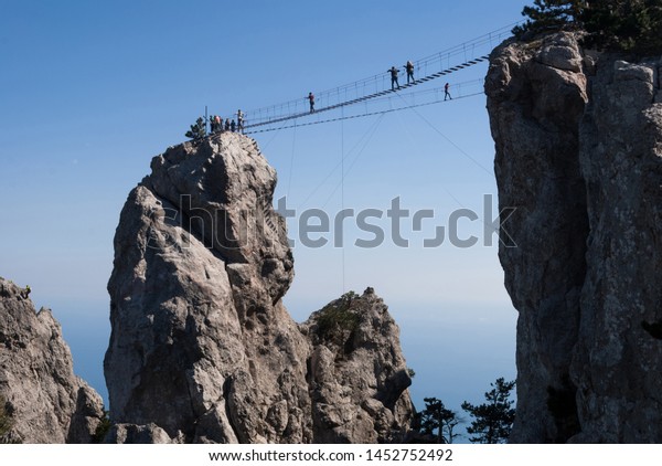 Tourist walking on a\
rope bridge on Mount Ai-Petri. Amazing view of the high rope bridge\
with traveler above abyss against the Black Sea coast. Scenery of\
Crimea in summer.