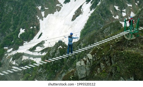 Tourist walks on rope suspension bridge on Rosa Peak (2320 m.) in Sochi, Russia. People on hanging footbridge above chasm, extreme attraction. Beautiful landscape, mountain peak in clouds and snow