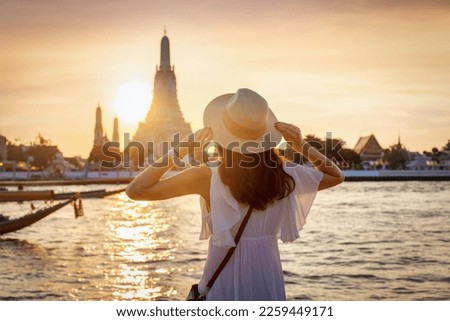 A tourist woman enjoys the view to the famous Wat Arun temple in Bangkok, Thailand, during golden sunset time