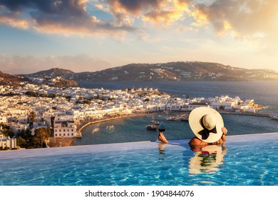 A Tourist Woman With A Glas Of Wine In A Swiming Pool Enjoys The View Over The Town Of Mykonos Island, Greece, During Summer Sunset Time