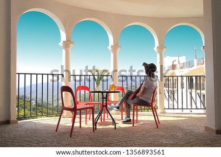 tourist woman with hat, with pants, sitting on the terrace of a bar drinking a beer, with some flowers, next to some arches and the turquoise sky, white village of Frigiliana, Malaga, Andalusia, Spain