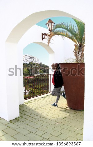 tourist woman with hat, with pants, standing, leaning on railing under arches and turquoise sky, white village of Frigiliana, Malaga, Andalusia, Spain,