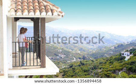 tourist woman with hat, with pants, standing, leaning on railing on a balcony under arches and the turquoise sky, white village of Frigiliana, Málaga, Andalusia, Spain,