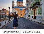 A tourist woman with hat walks down a road in the Vaporia district of Ermoupolis, Syros, Greece, during summer evening time
