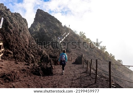 Tourist woman hiking along cloudy edge of active volcano crater of Mount Vesuvius, Province of Naples, Campania region, Italy, Europe, EU. Volcanic landscape full of stones, ashes and solidified lava
