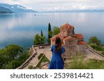 Tourist woman looking at iconic Church St. John at Kaneo. Panoramic view of calm Lake Ohrid, North Macedonia. Popular tourist attraction in Balkans. Summer travel destination. UNESCO World heritage