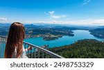 Tourist woman with panoramic view of idyllic lake Woerthersee in summer seen from observation tower Pyramidenkogel, Carinthia, Austria. Lakeside village surrounded by hills and mountains Austrian Alps