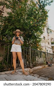 Tourist Woman Photographer Standing Under Pomegranate Tree In Old Greek City. Summer Vacation Concept. Lady Exploring Island. Amateur Taking Photos With Small Digital Camera.