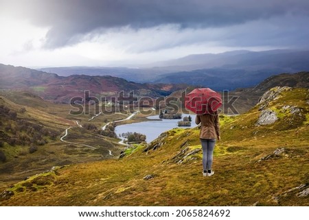 A tourist woman with a scotish pattern umbrella enjoys the view over the highlands to Lock Knokie in Scotland during autumn time