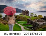A tourist woman with a scottisch pattern umbrella looks at the famous Urquhart Castle at Loch Ness during autumn time, Scotland