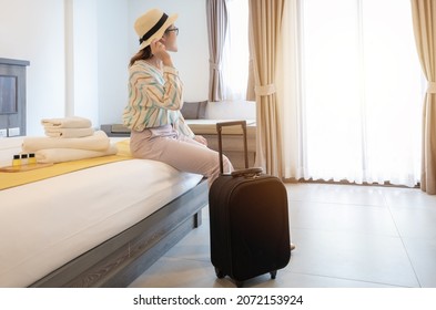 Tourist Woman Sitting On Bed And Looking To Beautiful View Outside The Room With Her Luggage In Hotel Bedroom After Check-in. Conceptual Of Travel And Accommodation.