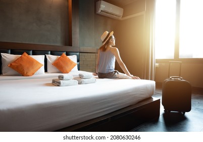 Tourist Woman Sitting On Bed With Her Luggage In Hotel Bedroom After Check-in And Looking To Beautiful View Outside Window. Conceptual Of Travel And Accommodation.