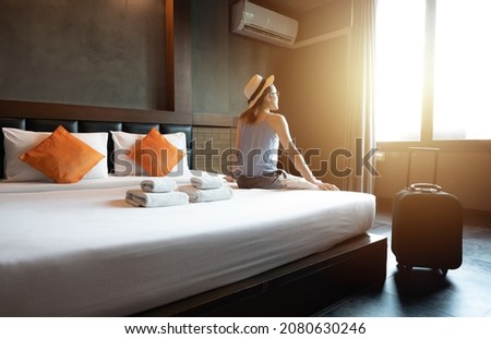 Tourist woman sitting on bed with her luggage in hotel bedroom after check-in and looking to beautiful view outside window. Conceptual of travel and accommodation.