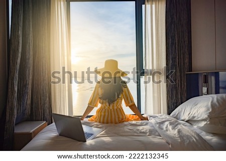 Tourist woman sitting on bed nearly window, looking to Beautiful sunset above sea or ocean, reflection of sun in the water after video conference meeting with colleagues , online working on laptop.  