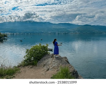 Tourist woman standing on edge of rock formation with panoramic morning views of Lake Ohrid, North Macedonia. Calm water surface reflecting surrounding hills and mountains. Summer travel destination - Powered by Shutterstock