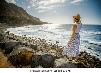 A tourist woman in a straw hat standing above El Socorro beach, admiring the landscape. The rocky shoreline, crashing waves, and cliffs evoke a sense of peace and natural beauty. - Powered by Shutterstock