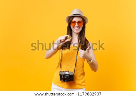 Tourist woman in summer casual clothes, hat holding bitcoin, metal coin of golden color isolated on yellow orange background. Female traveling abroad to travel on weekends getaway. Air flight concept