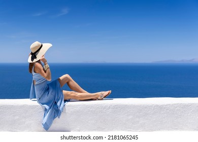 A tourist woman in a summer dress sits on a white wall on front of the blue, Aegean See of Greece, Cyclades island - Powered by Shutterstock