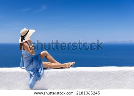 A tourist woman in a summer dress sits on a white wall on front of the blue, Aegean See of Greece, Cyclades island