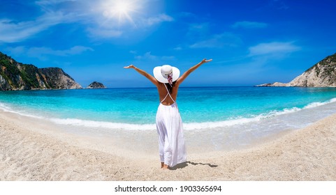 A Tourist Woman In White Dress And Hat Enjoys The Beautiful Beach Of Petani, Kefalonia Island, Greece, During Her Summer Holiday Time