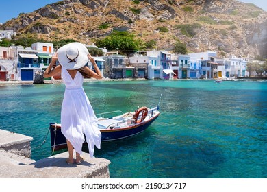 A tourist woman in a white dress and hat looks at the colorful fishing village of Klima on the island of Milos, Cyclades, Greece, during summer time - Shutterstock ID 2150134717