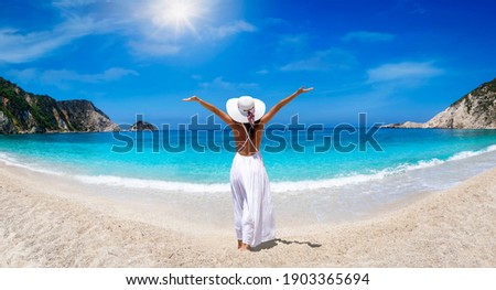 A tourist woman in white dress and hat enjoys the beautiful beach of Petani, Kefalonia island, Greece, during her summer holiday time
