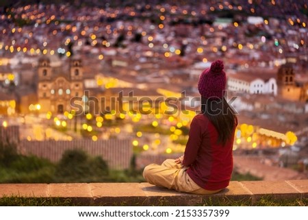 Tourist with woolen cap sitting half-sideways on San Cristobal viewpoint looking the illuminated main square of Cuzco at night