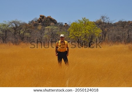Tourist in yellow shirt standing in field of tall yellow grass with Matobo hills in background in sunny day, Zimbabwe National Park